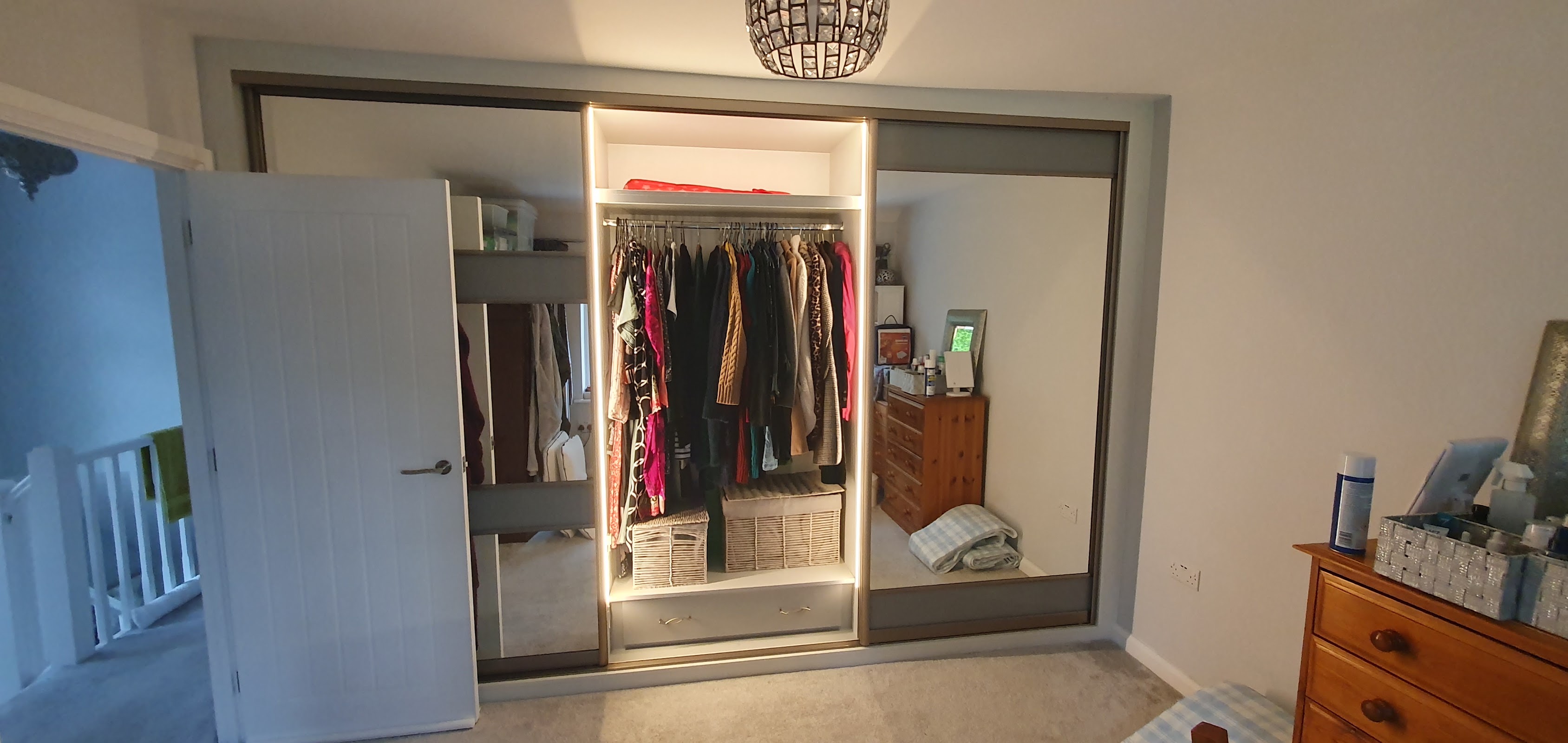 A three-panel sliding wardrobe in mirror and soft grey vinyl, with a lit central open section for hanging — mirrors bouncing light around a family bedroom.
