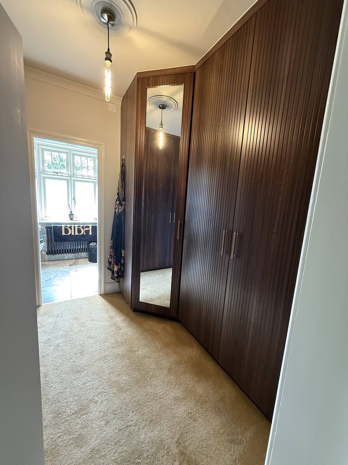 A warm walnut corner wardrobe run with a full-height mirror, wrapping a dressing area in a period Solihull home.